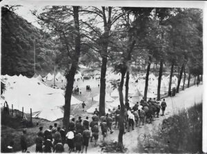 Historisches Zeltlager in einer bewaldeten Gegend, vermutlich aus den 1920er Jahren, mit Menschen, die zwischen Zelten und Bäumen umhergehen