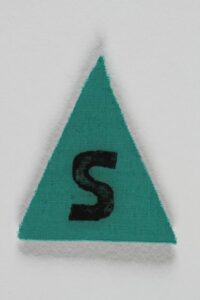 Ein grünes Dreieck mit einem "S" in der Mitte. Abzeichen der als “Berufsverbrecher” stigmatisierte Menschen auf den Häftlingsjacken tragen mussten.